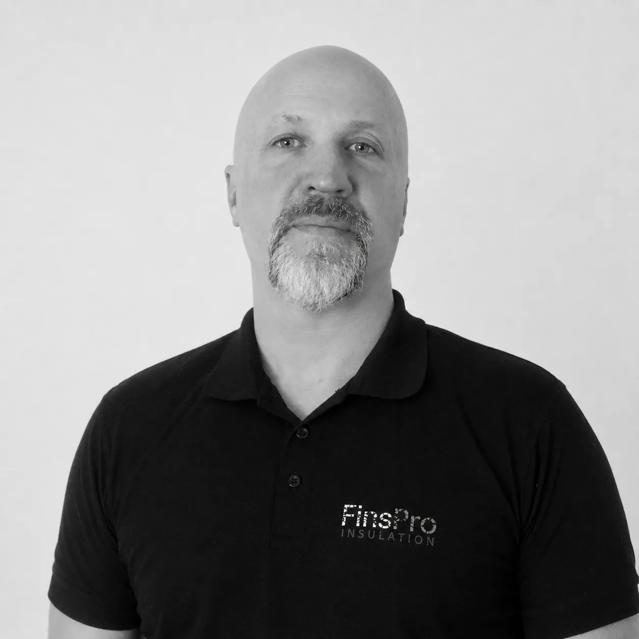Mustavalkoinen kuva kaljusta miehestä, jolla on harmaa pukinsaksa. Hänellä on yllään musta pikeepaita, jossa lukee "FinsPro INSULATION". Mies katsoo suoraan kameraan valkoista taustaa vasten.