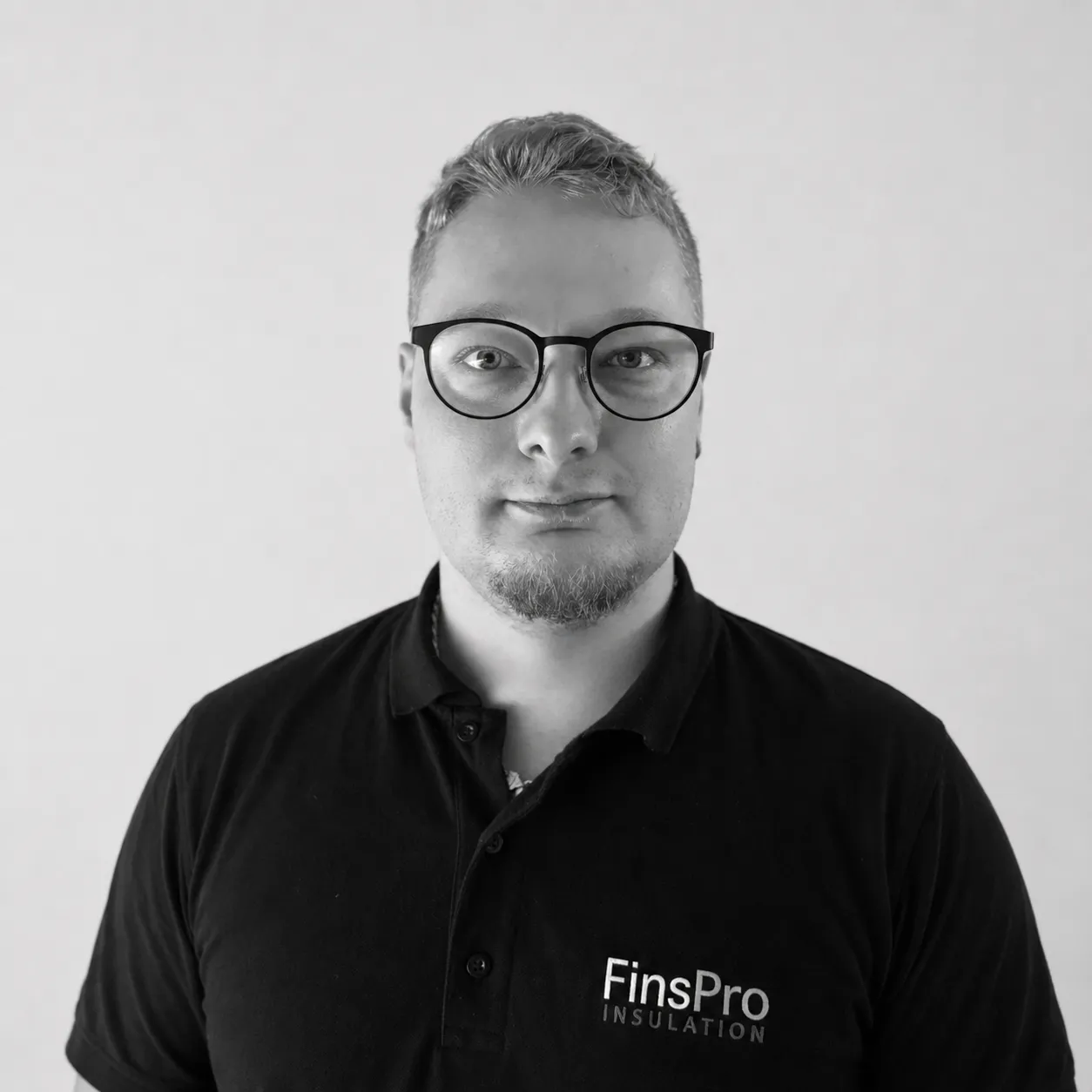 Mustavalkoinen muotokuva miehestä, jolla on vaaleat lyhyet hiukset, parranpätkä ja silmälasit. Hän katsoo suoraan kameraan. Yllään hänellä on tumma poolopaita, jossa lukee 'FinsPro INSULATION'. Tausta on vaalea ja tasainen.