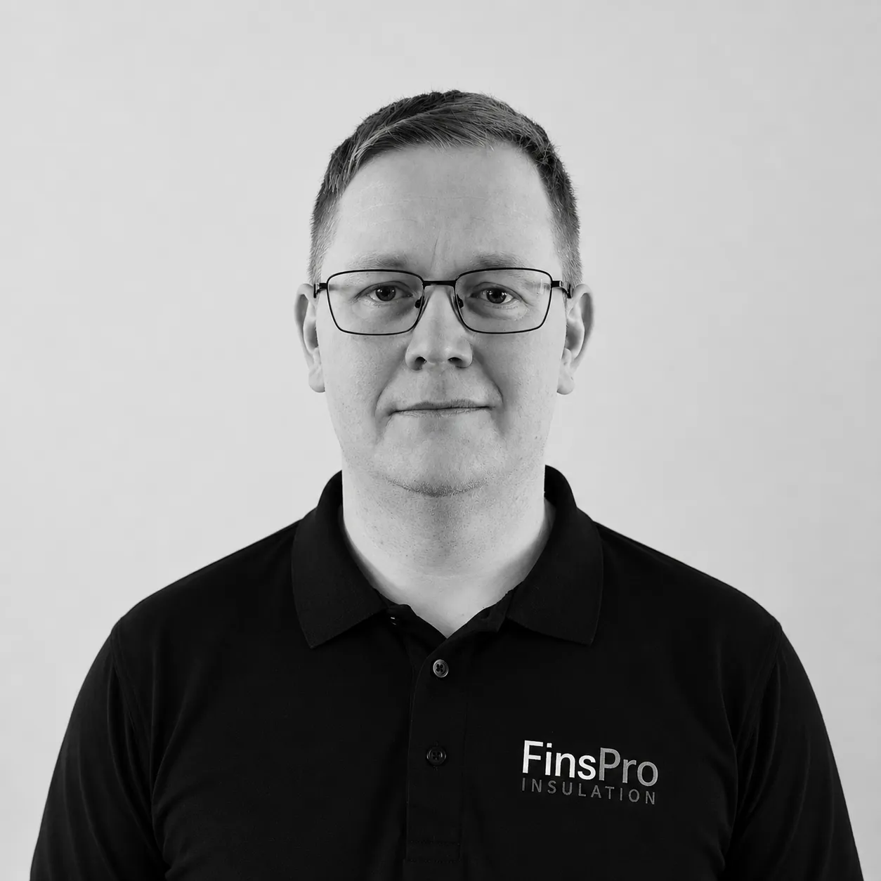Mustavalkoinen kuva miehestä, jolla on silmälasit ja musta pikeepaita. Paidan vasemmassa rinnassa on logo "FinsPro INSULATION". Mies katsoo suoraan kameraan vaaleaa taustaa vasten.